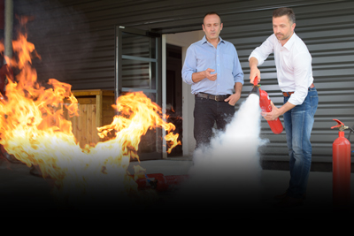 two men practicing fire extinguisher techniques on flames demonstrating fire safety training with eight different extinguishers available